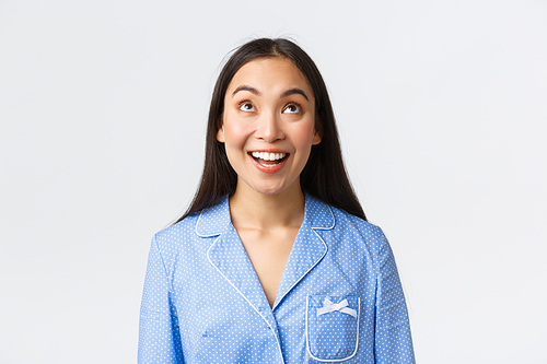 Close-up of amused and curious pretty asian woman in blue pajama spot interesting promo, looking up with broad happy smile, reading announcement with delighted face, white background.