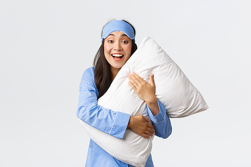 Smiling excited pretty asian girl in blue pajamas and sleeping mask, hugging soft comfortable pillow and looking surprised or amazed at camera at sleepover, white background.