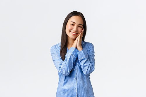 Dreamy smiling asian girl in blue pajamas getting ready sleep, leaning face on palms and grinning as lying bed. Charming korean female at bedtime standing over white background.