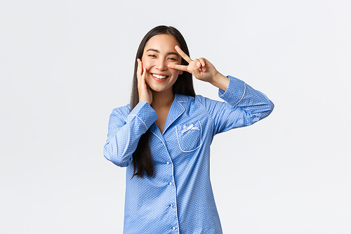 Charming silly asian girl in blue jammies showing peace sign and touching cheek kawaii, blushing and smiling, satisfied with clean perfect skin, use skincare products, white background.