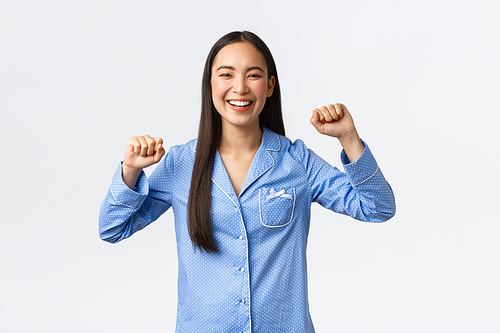 Enthusiastic happy asian girl in blue pajamas, smiling pleased and stretching hands after good sleep, feeling energized in morning, pumped up from great nap, white background.