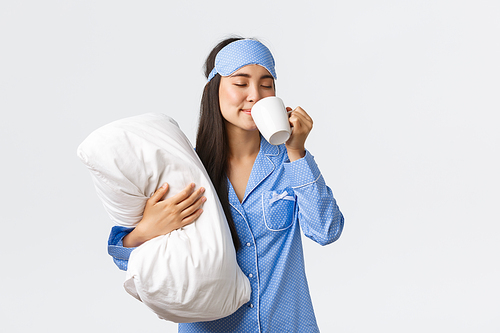 Morning lifestyle, breakfast and people concept. Smiling beautiful asian girl in pyjamas and sleeping mask, hugging pillow, having coffee in bed and watching tv, home leisure on weekends.