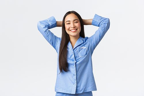 Smiling pleased and relaxed asian girl looking happy in blue pajamas, holding hands behind head as lying in bed satisfied, feeling relieved and delighted over white background.