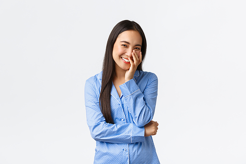Smiling beautiful korean girl in blue pajamas touching clean pure skin after applying skincare product, cleanser, laughing coquettish and blushing, feeling happy, white background.