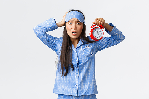 Woman having panic as showing alarm, being late for work, overslept to morning exam, holding hand on head and frowning nervous, standing in blue pajamas and sleeping mask anxious.