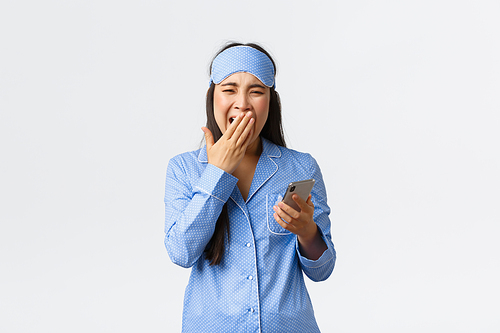 Technology, people and home leisure concept. Sleepy cute asian girl in pajamas and sleeping mask, waking up early, yawning exhausted and holding smartphone, turn-off mobile alarm.