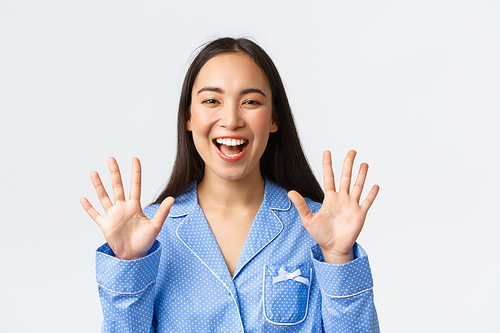 Close-up shot of happy attractive asian woman in blue pyjama showing ten fingers and smiling white teeth, explain main rules or making order, standing white background, recommend product.