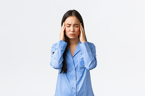 Asian girl in blue pajamas having insomnia, feeling unwell, touching temples grimacing from pain with closed eyes, suffer headache or migraine, need painkillers over white background.