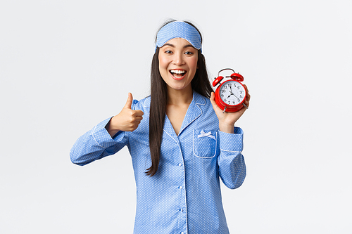 Enthusiastic and happy smiling asian girl in blue pajamas and sleeping mask, showing alarm clock and thumbs-up in approval, like waking up early for morning run, active and healthy lifestyle.