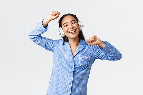 Home leisure, weekends and lifestyle concept. Cheerful happy asian girl in pajama having fun, dancing to music in earphones, listening favorite song on day-off, standing joyful white background.