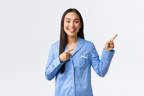 Smiling beautiful asian girl in blue pajamas making announcement, pointing fingers upper right corner to show big news or promo banner, recommend advertisement over white background.