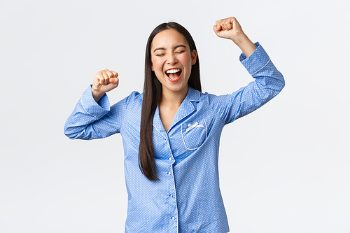 Cheerful triumphing asian woman in blue pajamas chanting, smiling and shouting yes delighted or relieved, raising hands up in success and victory, celebrating great news, white background.