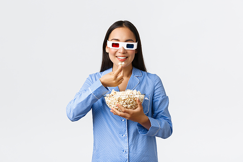 Home leisure, sleepover and slumber party concept. Interested pretty asian girl in blue pajamas and 3d glasses watching interesting movie, eating popcorn and smiling satisfied, white background.