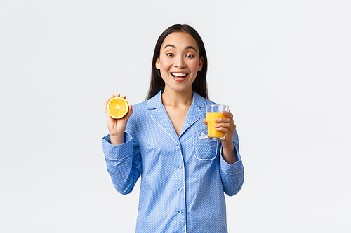Morning, active and healthy lifestyle and home concept. Smiling cheerful asian girl starting her day with fresh made orange guice, holding glass and half of orange, looking happy and energized.