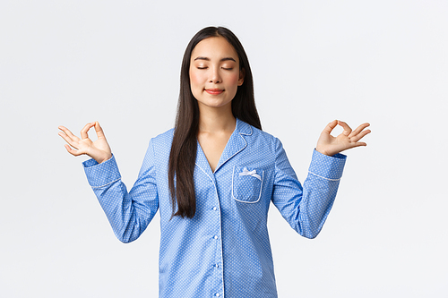 Calm and happy smiling asian girl in blue pajamas close eyes, meditating before sleep or in morning, looking relieved and peaceful, practice yoga meditation over white background.