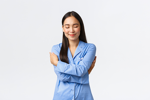 Happy dreamy tender asian girl in blue pajamas, close eyes and smiling as daydreaming, hugging herself, embracing own body in jammies, standing white background relaxed, daydreaming.