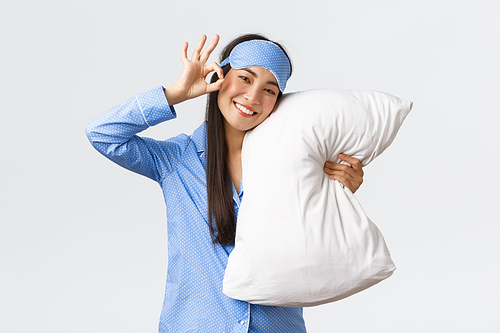 Satisfied happy smiling and cute asian girl in blue pyjama and sleeping eyemask, hugging pillow in mask, showing okay gesture, recommend product or medication for sleep, white background.