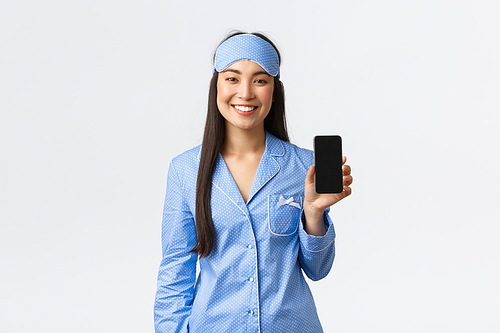 Technology, people and home leisure concept. Happy smiling asian girl in pajamas and sleeping mask tracking her sleep with mobile app, showing smartphone screen and look pleased, white background.
