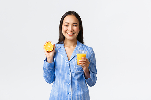 Morning, active and healthy lifestyle and home concept. Smiling cheerful asian girl starting her day with fresh made orange guice, holding glass and half of orange, looking happy and energized.