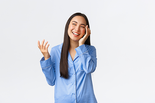 Beautiful asian girl getting ready sleep, touching soft clean face after applying skincare night products, wearing pajamas, laughing and looking upper left corner happy, white background.