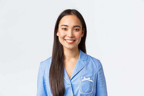 Close-up of good-looking asian girl in blue pajama with perfect white teeth, smiling at camera delighted, waking-up in morning enthusiastic, had good night sleep, standing white background.