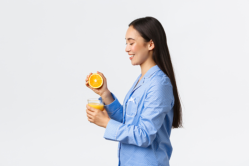 Morning, active and healthy lifestyle and home concept. Profile or beautiful healthy asian girl in blue pajamas squeezing orange juice in glass and smiling happy, starting day right.