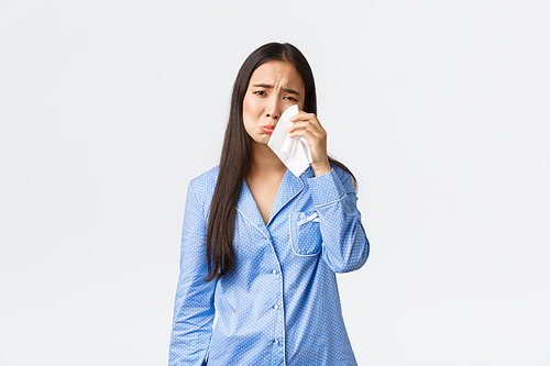 Distressed silly asian girl in blue pajamas feeling heartbroken, staying in bed in bad mood, wipe tears with tissue, sobbing and crying depressed, grieving over white background.