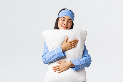 Beautiful smiling asian girl in sleeping mask and pajama, having sweet dream, hugging pillow with silly grin and closed eyes, standing over white background, unwilling wake-up.