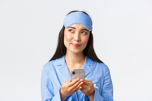 Close-up of thoughtful beautiful asian girl in blue pajama and sleeping mask having an idea, looking away with cunning smile as using mobile phone, texting in bed, standing white background.