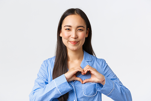 Close-up of lovely feminine asian girl in blue pajama express love and care, showing heart gesture, smiling happy as like something, standing white background delighted. Copy space