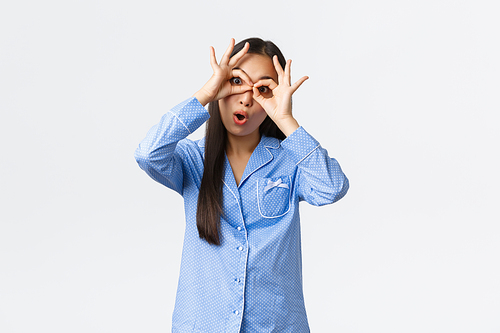 Funny and upbeat pretty asian girl in blue pajamas making faces, showing fake glasses with hands and playfully stare camera, having fun mocking someone at sleepover, white background.