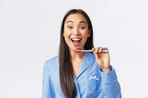 Cheerful pretty young asian girl in blue pajama waking-up, brushing teeth with broad enthusiastic smile, holding toothbrush near white teeth, white background. Copy space