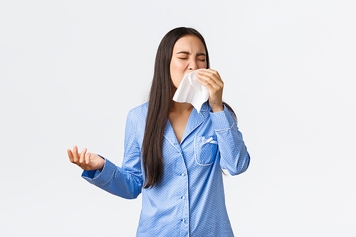 Asian girl in blue pajamas sneezing in napking, having seasonal allergy or got sick, staying home with runny nose, wearing jammies, feeling unwell in bed, standing white background.