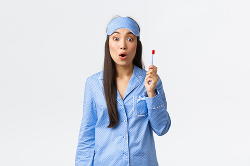Hygiene and people at home concept. Girl have great idea while brushing her teeth in bathroom before going sleep. Asian female in pajamas and sleeping mask holding toothbrush and look amazed.