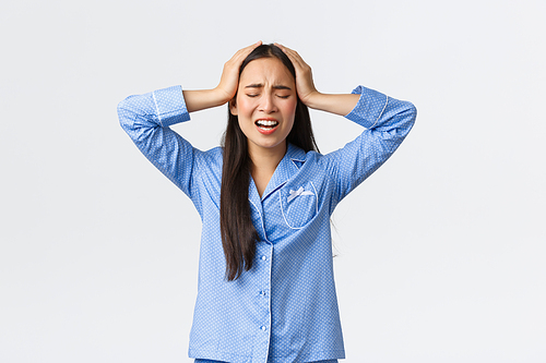 Troubled alarmed asian girl in blue pajamas standing desperate and anxious, holding hands on head in denial and shouting, feeling panic and frustration as having big problem, white background.