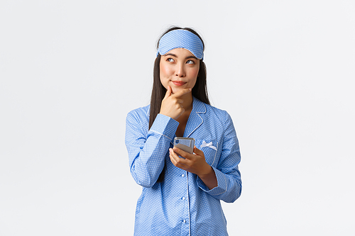 Technology, people and home leisure concept. Thoughtful and creative cute female blogger, asian girl in pajamas and sleeping mask have idea, thinking and holding mobile phone, creating post.