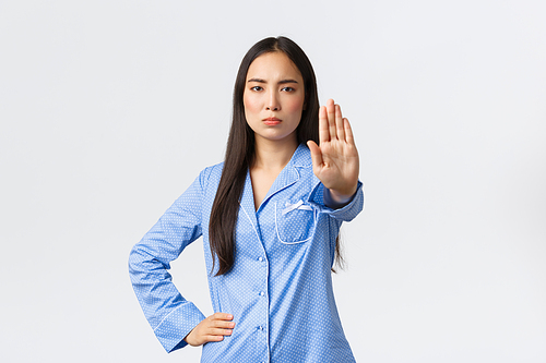 Serious-looking determined asian girl in blue pajamas extend hand to show stop, prohibit action, making warning and disapprove bad behaviour, forbid something over white background.