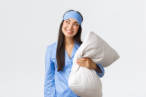 Cunning and thoughtful kawaii asian girl in blue pajama and sleeping mask, holding pillow and looking curious upper left corner, smiling sly as having idea, imaging something, white background.