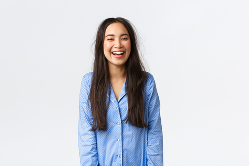 Smiling happy and energized asian girl with messy hair wearing pajamas and laughing, waking-up enthusiastic and cheerful, having good night sleep, standing white background.