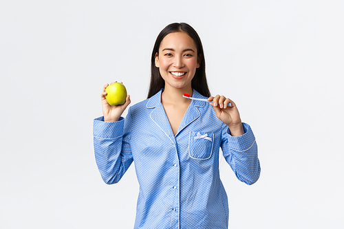 Morning, active and healthy lifestyle and home concept. Smiling beautiful asian girl showing toothbrush and green apple, have perfect white teeth and give advice on mouth hygiene, white background.
