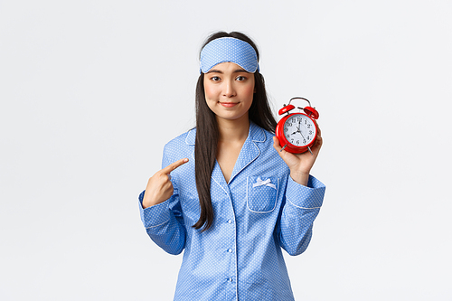 Oopsie, awkward silly girl in blue pajamas and sleeping mask pointing finger at alarm clock and smiing guilty as overslept or forgot set-up it, standing white background sorry.