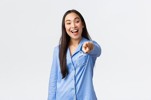 Cheerful beautiful asian girl in blue pajamas pointing finger at camera and smiling happy, picking or choosing you, making decision, being confident and standing over white background.
