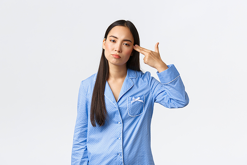 Annoyed and bothered asian girl in blue pajamas looking with reluctant, shooting herself with gun gesture as feeling fed up, tired of hearing or seeing something boring or dumb, white background.
