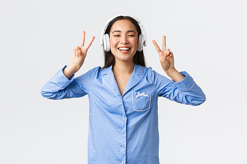 Home leisure, weekends and lifestyle concept. Kawaii asian girl in pajama listening music in wireless headphones and showing peace gesture with broad happy smile, white background.