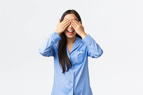 Upbeat excited smiling asian girl in blue pajamas, close eyes and grinning with anticipation as waiting for surprise gift, standing blindfolded delighted over white background.