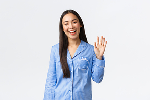 Beautiful asian woman in blue pajamas smiling happy and waving hand to say hello, greeting girlfriend and welcome to sleepover party, standing white background with hi gesture, white background.