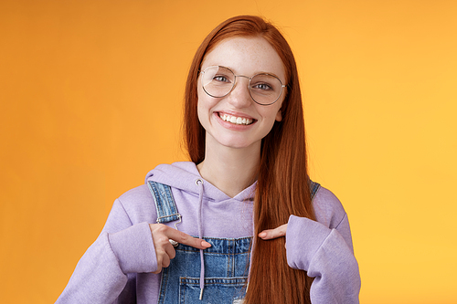 Happy lively friendly redhead european girl pointing herself suggesting help boasting telling own accomplishments smiling white teeth delighted volunteering, being picked, orange background.