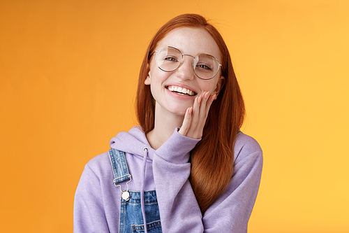 Charismatic happy tender redhead smiling european girl 20s glasses hoodie denim overalls having fun enjoying summer holidays chilly evening laughing joyfully touch pure clear skin, orange background.