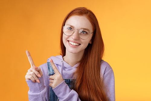 Carefree charismatic happy silly young cute redhead girl freckles blue eyes wearing glasses enjoy contemplating entertaining exhibition pointing looking left indicating, orange background.