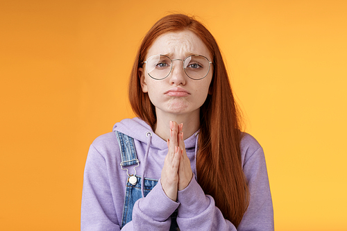 Please beg you. Clingy upset sad caucasian redhead girlfriend wearing glasses frowning grimacing pouting pleading help asking favour need urgently borrow something, orange background.